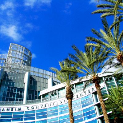 This image shows the exterior of the Anaheim Convention Center with its modern glass architecture and several palm trees against a vibrant blue sky.