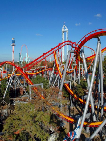 The image shows a large amusement park with multiple roller coasters and rides, featuring bright red and yellow tracks and lush greenery.