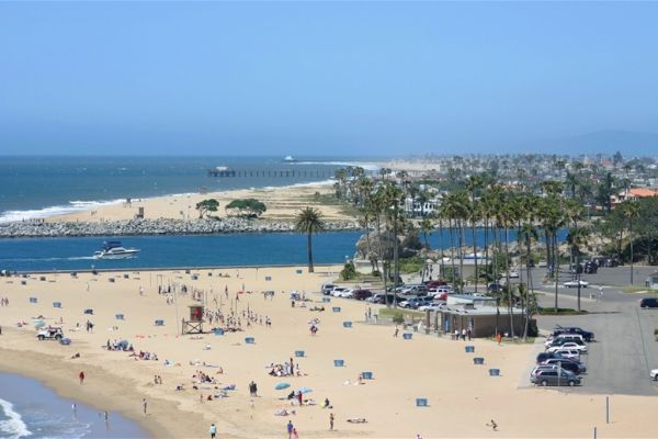 A scenic beach with people enjoying the sand and water, some cars parked, palm trees, and a clear blue sky in the background, ending the sentence.