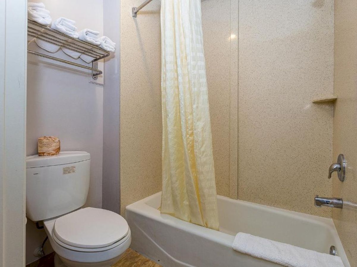 A bathroom with a toilet, bathtub, shower curtain, and rack holding towels. A small shelf above the toilet holds more towels, ending the sentence.