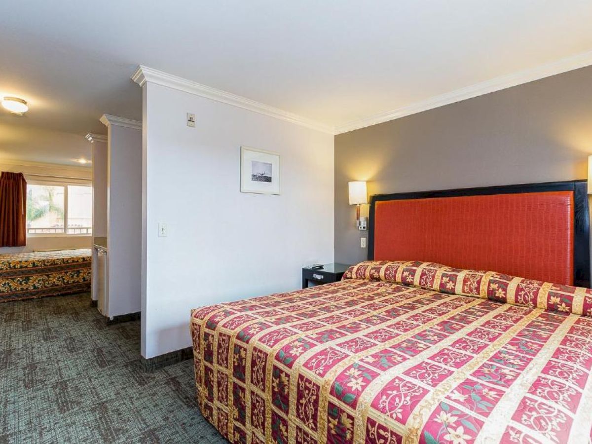The image shows a hotel room with a large bed, red and gold bedspreads, carpeted floor, wall lamps, and a separate area with another bed visible.