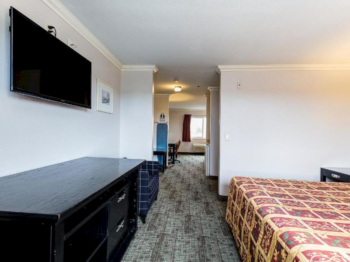 The image shows a hotel room with a bed, a TV mounted on the wall, a dresser, and a hallway leading to another section with additional furniture.