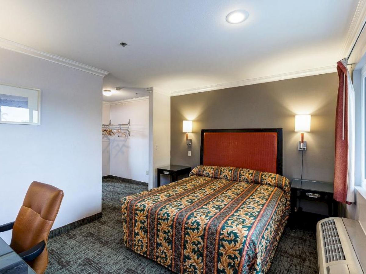 A hotel room with a patterned bed, two bedside tables, wall lamps, a desk with a chair, a window, and an open closet.