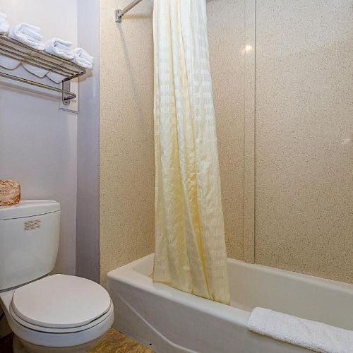 This image shows a small bathroom with a toilet, bathtub-shower combo, and towel rack holding folded towels. The decor is simple and clean.