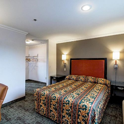 This image shows a hotel room with a bed, patterned bedspread, bedside lamps, desk, chair, and an air conditioning unit near the window.