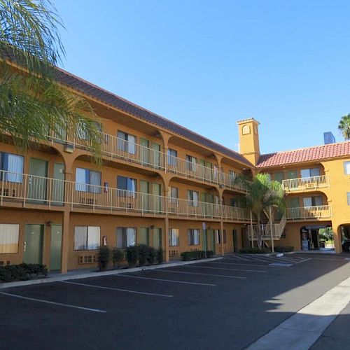 The image shows a three-story motel with a yellow exterior, an open parking area, and palm trees near the entrance on a clear day.