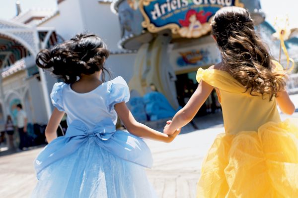 Two children in princess dresses are holding hands and walking towards an attraction called 