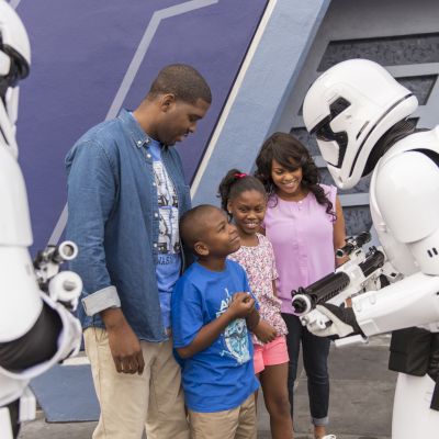 A family interacts with Stormtroopers in white armor and helmets at an amusement park or themed event, with everyone appearing to be happy.